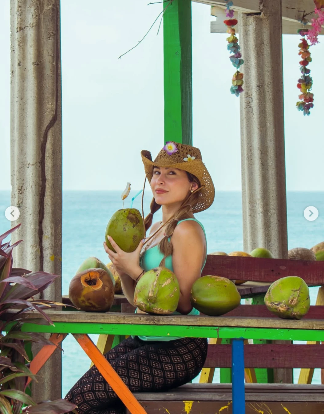 Andrea sentada en un kiosco de playa frente al mar, con sombrero de paja y un coco en la mano.